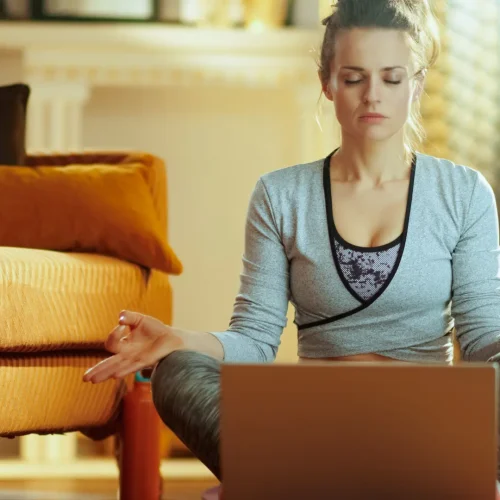 Frau in einem Wohnzimmer in Meditationshaltung vor einem Laptop am Meditieren.